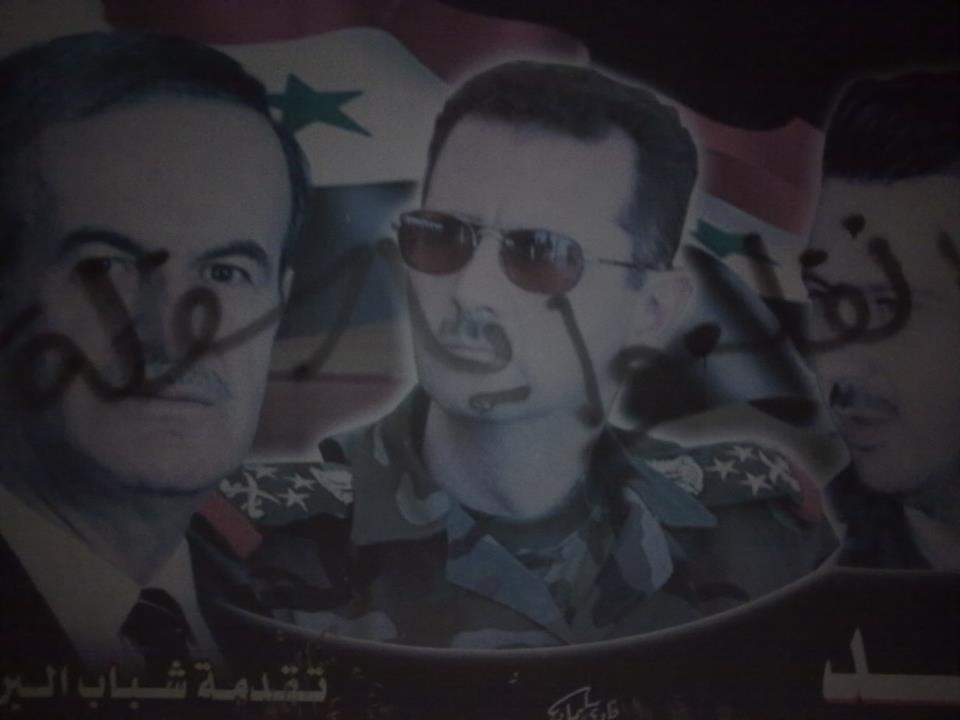 [Photo: One of Ayman's graffiti on a poster that depicts the Asad family in rural Jablah. It reads “Get Out You Varmints” (SyriaUntold)].