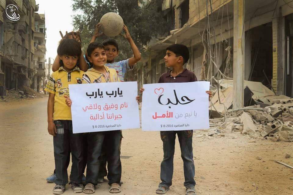 [Photo: Children in besieged Daraya (Rif Dimashq governorate) holding banners that celebrate the partial lifting of the siege of Aleppo - 8-8-2016 ( Local Council of Daraya Facebook page)].